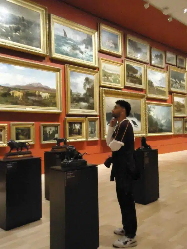 Alex Moulsdale Exercise Physiologist, standing, deep in thought while looking at a wall of framed oil paintings.