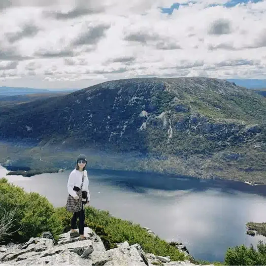 Ruri Zhang, Senior Physiotherapist, is standing on a rocky mountain summit overlooking an alpine lake and distant mountains.