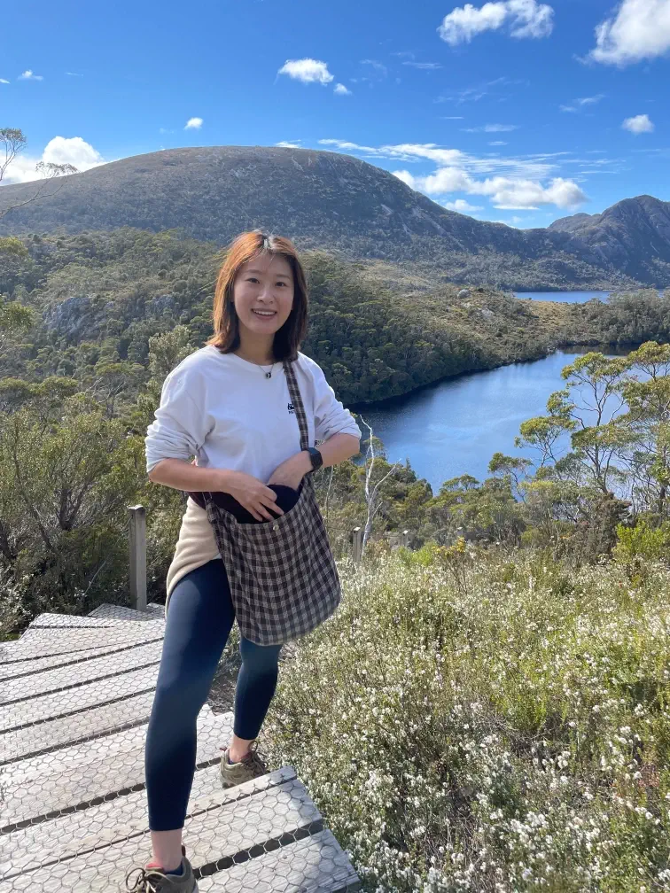 Ruri Zhang, Senior Physiotherapist, is standing on a wooden boardwalk high on a mountain overlooking a scenic lake and heavily forested valley on a sunny day.