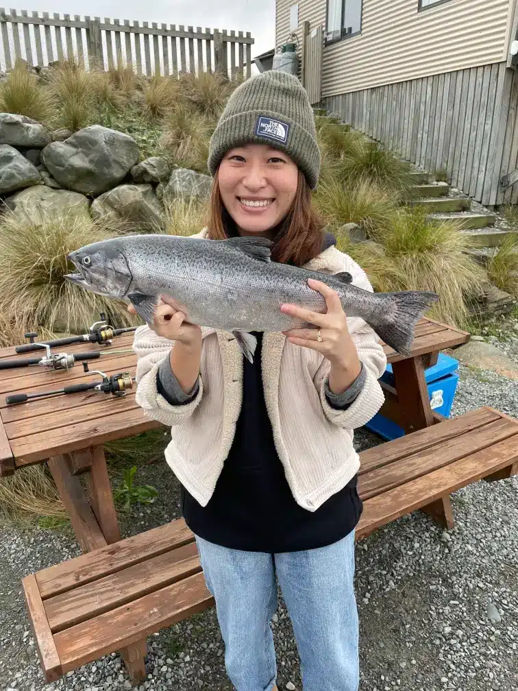 Ruri Zhang, Senior Physiotherapist, is proudly holding a large, freshly caught salmon while standing next to a wooden picnic table with fishing rods.