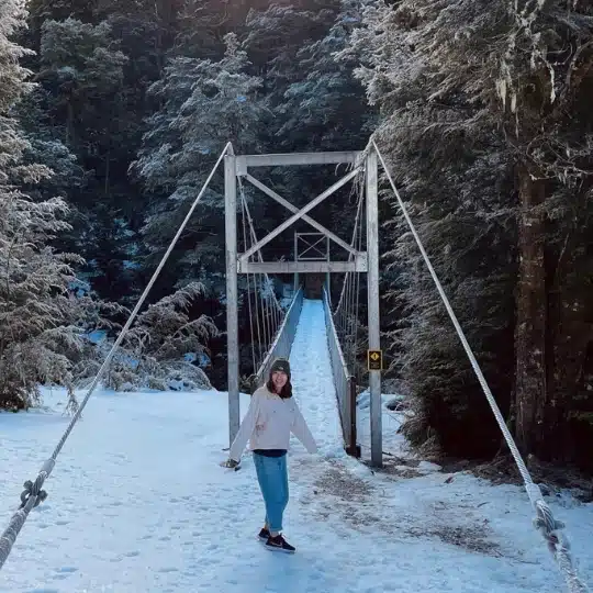 Ruri Zhang, Senior Physiotherapist, stands on a snowy path leading to a wooden suspension bridge in a forest of frost-covered trees.